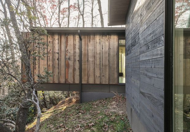 Exterior architectural view of a modern, elevated timber-clad dwelling set among mature woodland. The composition shows a dark horizontal weatherboard wall in the foreground, a projecting upper volume with vertically arranged weathered timber boards, narrow glazing, and a low flat roof. The building appears carefully integrated into a sloping, leafy site.