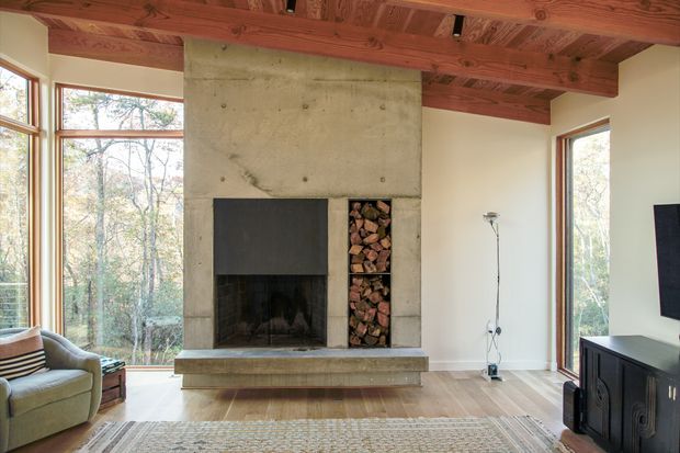 A warm, light-filled living room centred on a large exposed concrete fireplace with an integrated firewood niche. Full-height corner glazing looks into a wooded setting, while exposed timber ceiling beams, pale walls, timber flooring, and a dark built-in media cabinet define the room. A lounge chair, floor lamp, television, and rug furnish the space.