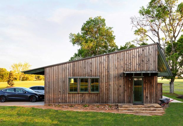 Single-storey timber-clad building viewed from the exterior, with a simple sloping gable-like roof form, dark-framed windows and entry door, and a small stepped entrance. Vehicles are parked beside the building, with open lawn and mature trees surrounding the site.