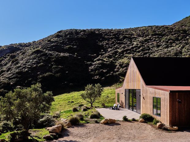 Exterior view of a compact contemporary timber-clad building set against a steep, rugged vegetated hillside. The structure has a distinctive steep triangular gable, low adjoining roof volume, large dark-framed glazed doors and windows, and a simple paved terrace surrounded by native-looking landscaping and exposed earth.