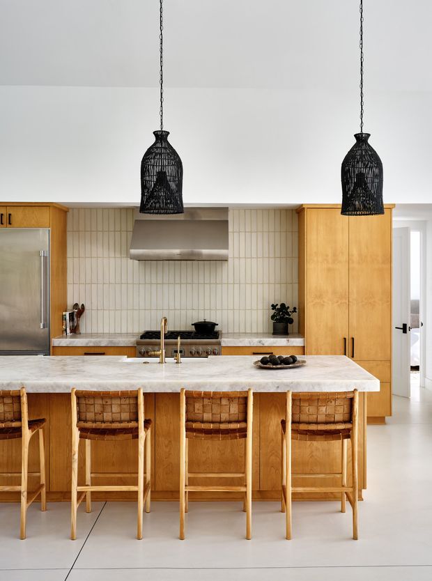 A bright, contemporary kitchen centred on a large white marble island with a timber-clad base and four woven timber bar stools. Warm natural timber cabinetry surrounds a pale vertical tile splashback, stainless-steel appliances, and a simple integrated rangehood. Two black woven pendant lights hang above the island, while a doorway opens to an adjacent bright space.
