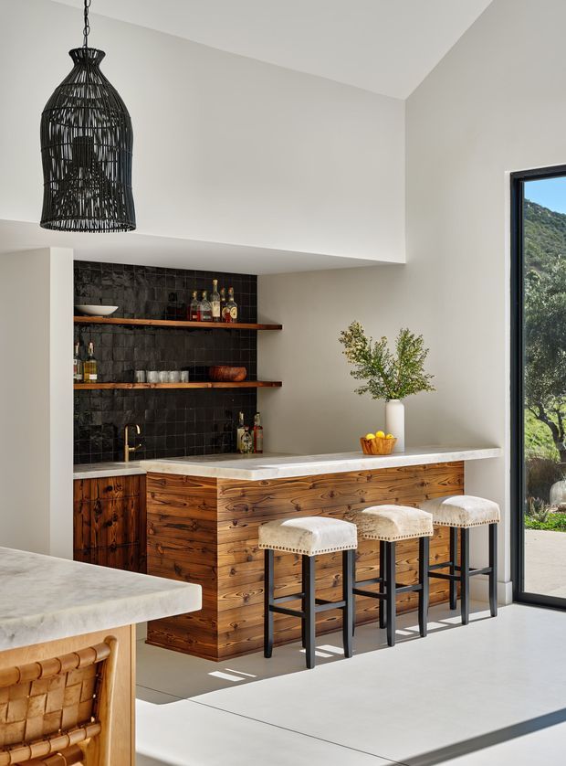 A refined open-plan home bar and entertaining area with a timber-fronted island, pale stone benchtop, three upholstered bar stools, dark textured tile splashback, and open timber shelves displaying bottles and glassware. A large black woven pendant light hangs above, while full-height glazing provides views to the landscaped exterior. The space has a warm, contemporary rustic character with strong natural timber detailing.