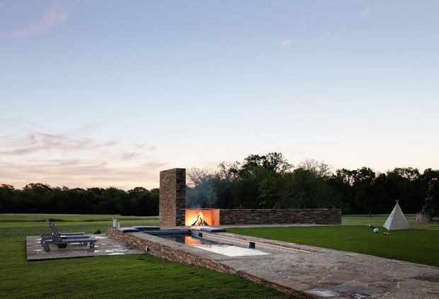 A landscaped outdoor entertainment area with a long rectangular swimming pool, paved stone terrace, lounge chairs, and a linear outdoor fireplace integrated into a low stone wall. The fireplace has a tall stone chimney and a visible fire, with open lawn and mature trees beyond. A small tent-like structure is visible at the far right.
