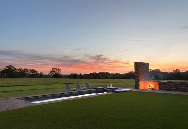 A landscaped outdoor entertaining area at sunset, featuring a long narrow reflecting pool or swimming pool, large manicured lawn, dark lounge chairs, and a contemporary masonry outdoor fireplace with a glowing fire and low stone wall. The open rural-looking setting and dramatic sunset create a calm, refined atmosphere.