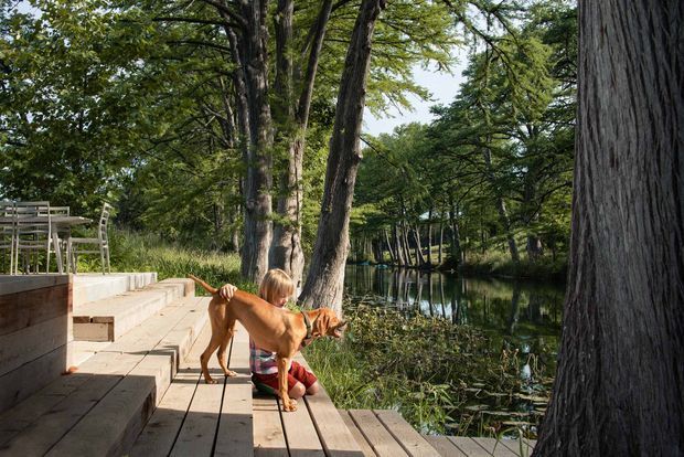 A timber deck and stepped outdoor terrace beside a calm, tree-lined pond or creek. A person sits on the deck with a dog, while mature trees and dense greenery frame the water. The setting feels natural, shaded and relaxed, with built timber decking and pale masonry steps integrated into the landscape.