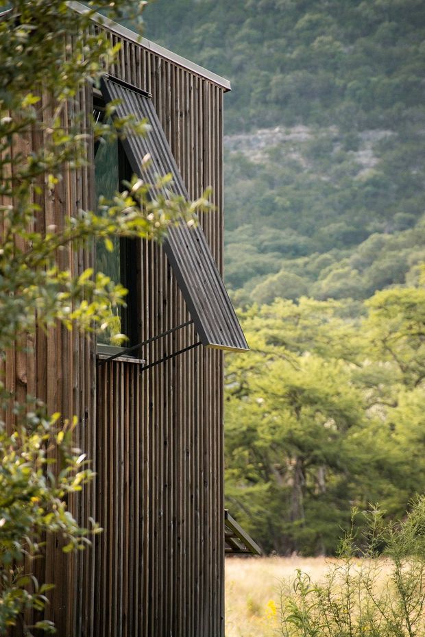 A close exterior view of a narrow, multi-level building clad in vertical dark timber battens. A projecting timber shutter or awning is opened beside a window, with a sloping roof edge visible above. The structure sits in a natural, wooded mountainous setting with vegetation in the foreground and background.