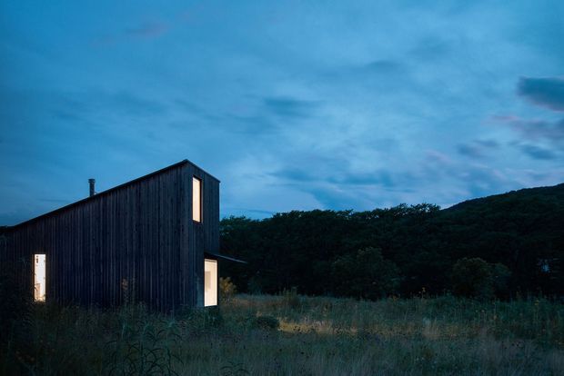 A compact, single-storey dark timber-clad building sits in a grassy rural clearing at dusk. The simple elongated form has a steeply sloping roof, minimal detailing, and several narrow illuminated openings, with wooded hills forming the background.