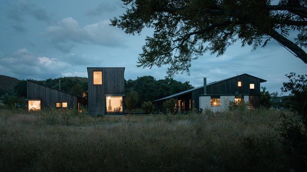 Twilight exterior view of a group of contemporary detached buildings set within a natural, overgrown landscape. The structures feature dark vertical timber cladding, simple pitched and shed roof forms, and warmly illuminated windows. A large tree frames the upper right of the image, while the subdued blue evening light creates a quiet rural atmosphere.