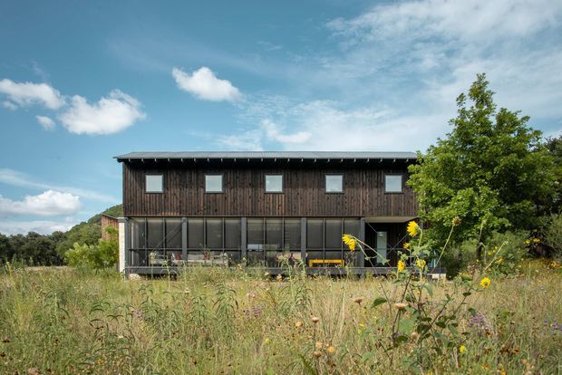 Exterior view of a simple two-storey rectangular building set within a meadow. The upper level has dark vertical timber cladding and a row of small square windows beneath a shallow pitched or nearly flat roof, while the lower level features extensive dark-framed glazing and a sheltered terrace or veranda. Mature vegetation and wildflowers surround the structure under a bright blue sky.