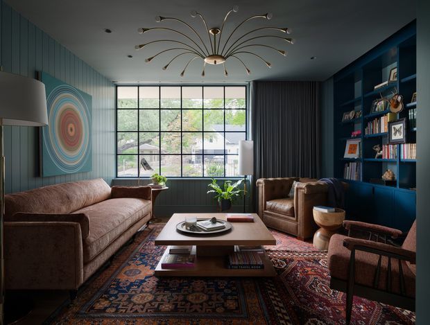 A richly styled living room with deep teal vertical wall panelling, a large black-framed gridded window, dark built-in shelving, and a sculptural mid-century-inspired branching chandelier. The room contains a brown upholstered sofa, leather armchairs, a low timber coffee table, patterned Persian-style rugs, and curated shelving. The palette is warm and moody with strong teal, brown, rust, and ochre accents, while daylight enters through the expansive window.