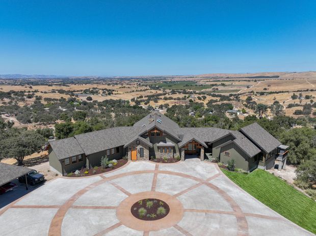 Aerial view of a large, low-rise lodge-style residence set on an expansive rural property. The building has multiple dark grey pitched and gabled roofs, dark exterior walls with lighter timber accents, and a broad circular concrete driveway or forecourt with radial terracotta-coloured paving bands and a central planted island. Rolling dry agricultural landscape and scattered trees extend into the distance.