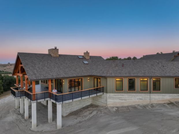 Exterior elevated view of a sprawling single-storey house at dusk, featuring a broad pitched shingle roof with multiple stone chimneys and skylights, muted green-grey vertical cladding, large dark-framed windows, and a covered timber porch opening onto a deck with black railings. The house sits on a substantial exposed concrete foundation over a bare, sloping site.