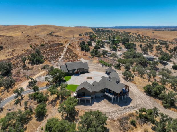 Aerial view of a large residential property set in a dry, rolling rural landscape. The multi-wing house has dark pitched roofs, covered verandas, and a broad paved driveway or courtyard, with detached outbuildings and scattered mature trees surrounding the site.