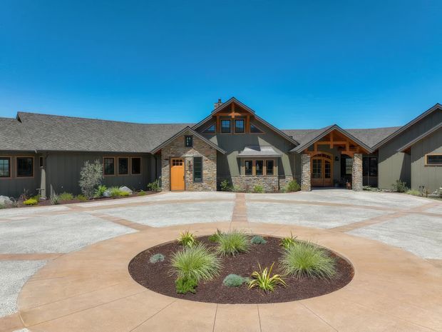 A broad single-storey residential building with multiple connected gabled roof volumes, dark grey exterior walls, natural stone veneer, timber gable detailing, and several timber-framed entrances. A large circular paved driveway or forecourt with a landscaped central planting bed dominates the foreground. The architecture has a lodge-like, rustic character with low-pitched shingle roofs and prominent timber accents.