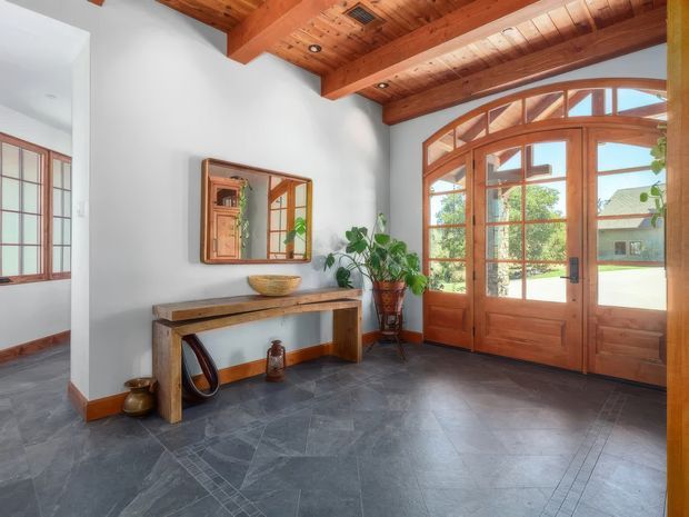 A bright, spacious entry hall with a large timber-framed glazed front door and arched transom, matching timber-framed sidelights, exposed timber ceiling beams and boards, pale plaster walls, and dark slate flooring. A simple timber console, wall mirror, woven bowl, and potted plants furnish the space. An adjacent room is visible through the opening on the left.