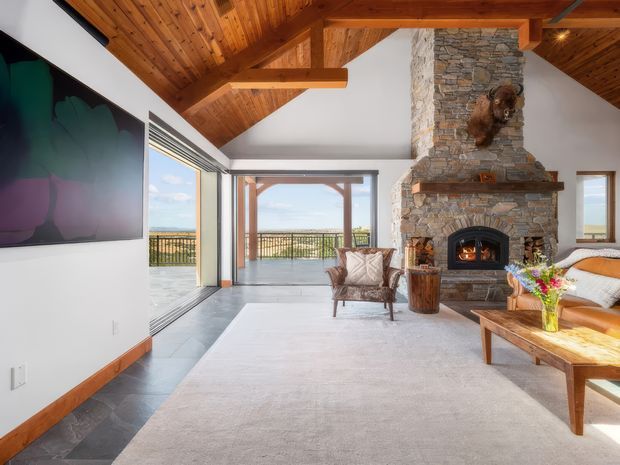 A spacious rustic living room with a dramatic open timber ceiling and exposed beams, large glazed doors opening to a balcony, and a substantial natural-stone fireplace with a wood-burning fire. The room includes a light sectional sofa, lounge chair, timber coffee table, pale rug, and colourful floral arrangement. Mountain or coastal scenery is visible beyond the glazing.