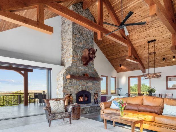 A spacious rustic living room with a dramatic exposed timber truss and vaulted timber-lined ceiling. A tall natural-stone fireplace with a timber mantel dominates the room, complemented by a leather sofa, upholstered armchair, timber coffee table, and contemporary ceiling fan. Large glazed openings connect the living area to a balcony or terrace with expansive outdoor views. The room has a warm, lodge-like atmosphere with natural materials and abundant daylight.
