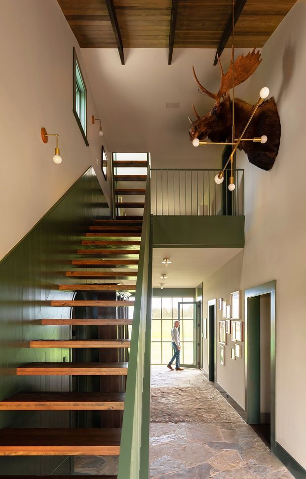 A tall, dramatic interior hallway centred on an open-riser staircase with thick dark timber treads and green painted metal or panelled sides. The stair rises toward a mezzanine with slender metal balustrading, while a long corridor extends toward full-height glazing at the far end. The space has a warm timber ceiling with exposed dark beams, pale plaster walls, green accents, stone flagstone flooring, sculptural wall lighting, and a prominent mounted moose head. Framed artwork lines the corridor walls.