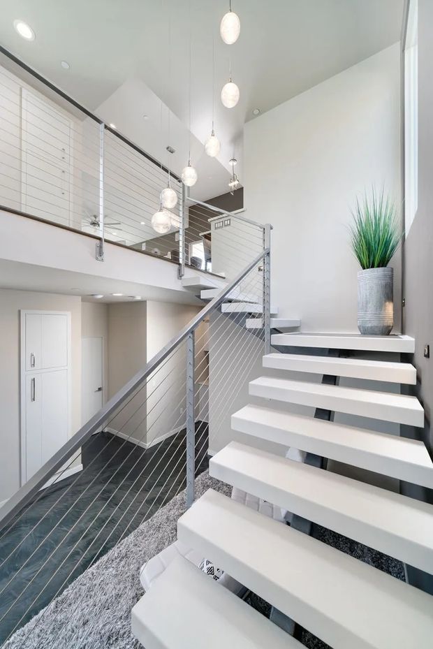 Bright contemporary multi-level stairway and landing with floating white stair treads, grey metal cable balustrades, dark flooring, and a tall open void. Multiple pendant lights hang through the stairwell, while white horizontal wall panelling and a potted plant add visual detail. A lower-level entry or hallway with built-in storage is visible beneath the mezzanine.