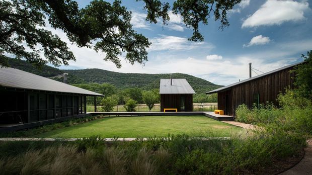 A low, single-storey contemporary compound arranged around a rectangular central lawn or courtyard. Dark timber-clad buildings with simple pitched metal roofs frame the view, with covered verandah-like glazing on the left, a smaller central outbuilding, and a larger building on the right. Native grasses and mature trees landscape the foreground, while wooded hills form the distant backdrop under a partly cloudy sky.
