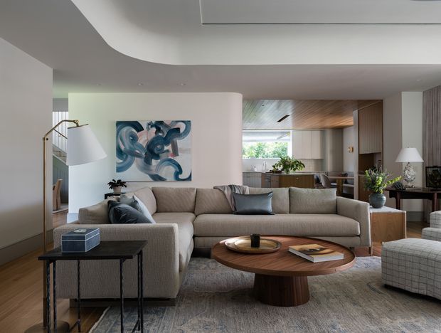 Contemporary open-plan living room with a large neutral upholstered sectional sofa, sculptural round timber coffee table, patterned rug, side tables, and abstract artwork. The room opens toward a visible kitchen and dining area, with warm timber ceiling and cabinetry, extensive glazing, and soft recessed lighting. A sculptural staircase and mixed neutral finishes contribute to a refined, welcoming interior.