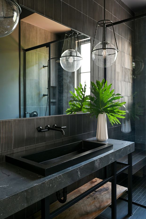 A dramatic, dark-toned contemporary bathroom vanity with a long black stone benchtop, integrated rectangular basin, wall-mounted matte-black tapware, dark vertically ribbed wall tiles, and a large mirror. A black-framed glass shower is visible in the background. Three suspended glass globe pendant lights and a tall white vase with green foliage add contrast. Open black metal vanity framing and timber shelving sit below the benchtop.