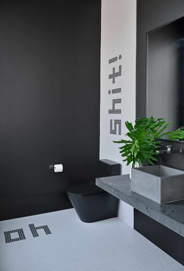 A dramatic contemporary bathroom featuring matte black walls, a black wall-hung toilet, a grey concrete-look vanity benchtop with a rectangular concrete basin, and a large mirror. White small-format mosaic tile flooring and a vertical white mosaic-tile feature strip provide contrast, with black pixel-style lettering and symbols integrated into the tiled surfaces. A green plant adds a natural accent.