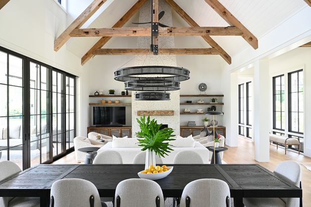 Bright open-plan interior showing a dining area in the foreground and a living room beyond. A long dark timber dining table with upholstered chairs faces a white stone fireplace and built-in shelving. Large black-framed glazed doors and windows line the sides, while exposed timber trusses and a vaulted white ceiling create a spacious barn-like character. A contemporary circular metal chandelier hangs above the living area.