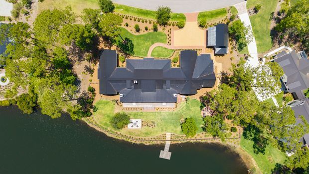 Aerial top-down view of a large residential property beside a waterfront, showing a dark-roofed multi-wing house, landscaped grounds, curved garden paths, an outbuilding, driveway, neighbouring homes, and a small private dock extending into the water.