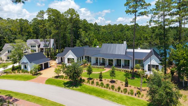 Aerial view of a large single-storey residential property set among mature trees beside a visible body of water. The home has multiple connected wings with dark grey pitched metal roofs, light-coloured exterior walls, extensive glazing, landscaped gardens, and a detached garage or outbuilding near the driveway.