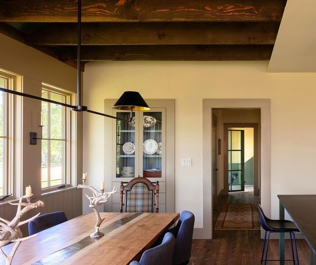 Warm, rustic dining area with a long timber dining table and dark upholstered chairs, positioned beside tall windows. A dark timber ceiling with exposed beams, cream walls, a glass-front display cabinet, and a black pendant light create a refined traditional farmhouse atmosphere. An open doorway leads to a hallway and glazed interior door beyond.