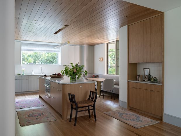 A bright open-plan kitchen and dining area with a central timber island, integrated pale cabinetry, timber flooring, and a warm timber-lined ceiling. Floor-to-ceiling built-in joinery frames a recessed display area on the right, while a dining nook and large windows bring in natural light. The space combines clean contemporary planning with warm mid-century-inspired timber detailing and furnishings.