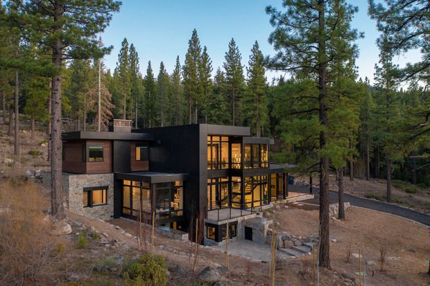 Exterior view of a contemporary multi-level house set among a dense pine forest on a sloping, rocky site. The building has a dark rectilinear facade, broad floor-to-ceiling glazing, projecting flat roof forms, and warm interior lighting visible through the windows. A paved driveway and stepped entry terrace are visible around the structure.