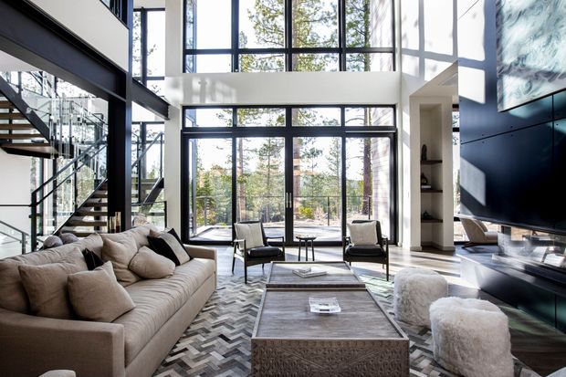 Bright, double-height contemporary living room with expansive black-framed floor-to-ceiling glazing overlooking trees. The space features a large neutral sectional sofa, sculptural lounge chairs, layered coffee tables, a chevron-patterned rug, open-riser staircase with glass balustrade, built-in shelving, and a large wall-mounted television with dark built-in cabinetry.