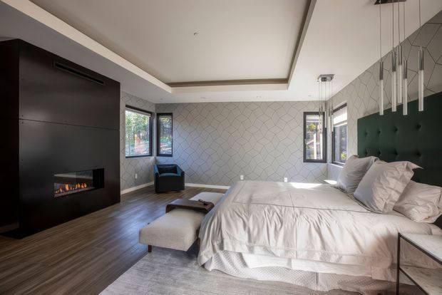 Contemporary bedroom with a large upholstered bed, dark green tufted headboard, white bedding, and a black feature wall incorporating a linear fireplace. The room has dark timber flooring, a pale geometric feature wall, several windows, a recessed tray ceiling, and sculptural pendant lights suspended above the bed. A soft grey rug and upholstered bench add texture to the otherwise refined, modern interior.