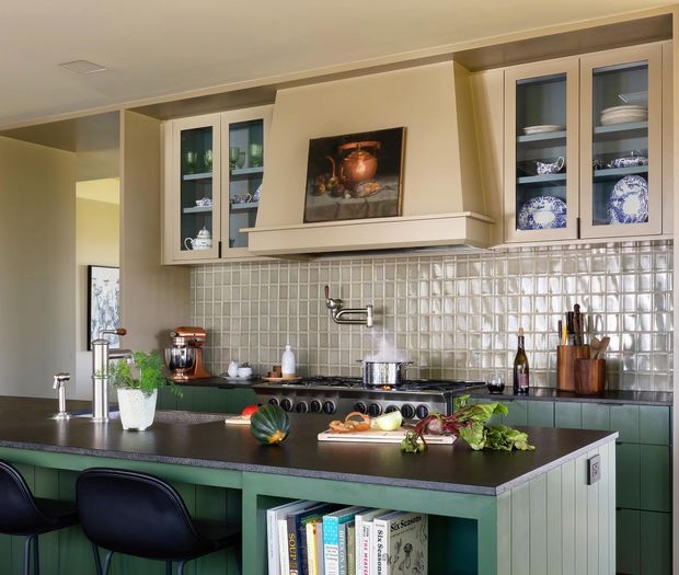 A well-appointed traditional kitchen with a large dark countertop island, green-painted base cabinetry, glass-front upper cabinets displaying crockery, and a full-height glossy square-tile backsplash. A classic range cooker sits beneath a prominent mantel-style rangehood surround, with open shelving and decorative accessories adding warmth.