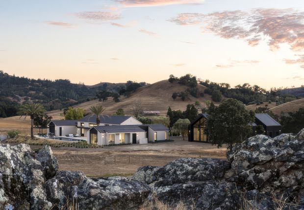 A contemporary rural property viewed from a rocky foreground, with low-profile light-coloured buildings, dark pitched roofs, extensive glazing, and a separate dark outbuilding set within rolling dry hills and scattered trees at sunset.