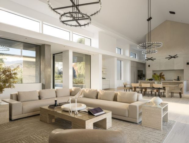 A bright, spacious open-plan living area with a large cream sectional sofa, textured neutral rug, substantial timber coffee table, and minimalist stone side table. The room opens directly into a contemporary dining and kitchen area, with floor-to-ceiling glazing, clerestory windows, high ceilings, and several sculptural circular pendant lights. The palette is soft beige, cream, white, and light timber, creating a refined and airy atmosphere.