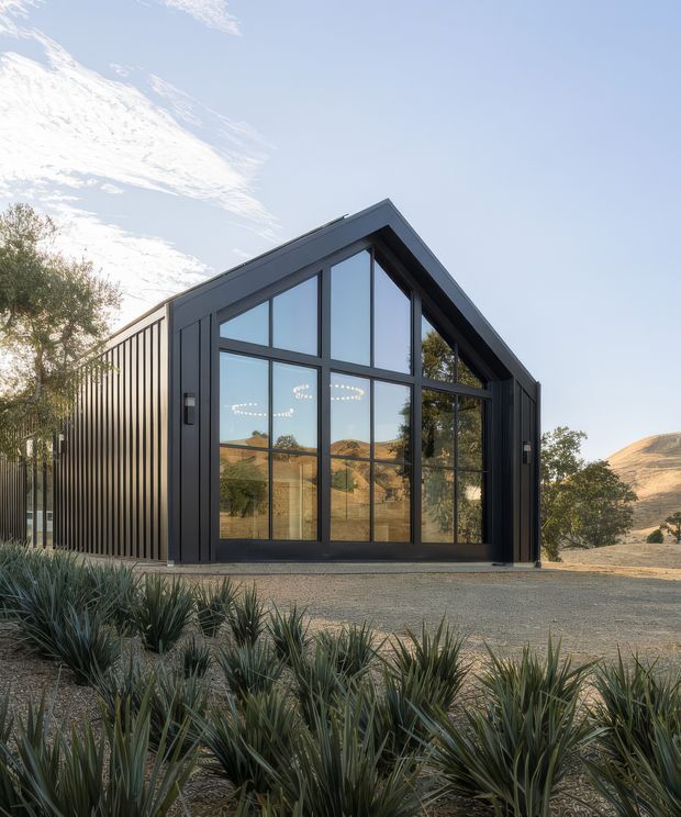 Exterior view of a compact contemporary barn-like building set in a dry, hilly landscape. The structure has a steep gable roof, dark vertical metal cladding, and a prominent floor-to-ceiling glazed gable facade with dark frames. Gravel surrounds the building, with drought-tolerant spiky planting in the foreground and trees and rolling hills visible around it.