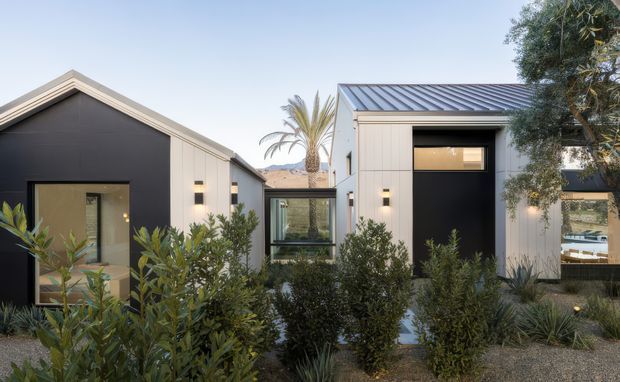 Contemporary single-storey residential buildings arranged around a landscaped courtyard. The structures feature simple gabled and shed-like roof forms, dark standing-seam metal roofing, light rendered or panelled walls, dark feature panels, and large glazed openings. Native-style planting and gravel landscaping surround the central glazed link and entrance areas.