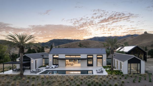A contemporary single-storey residence set in a dry, hilly landscape at sunset. The composition shows a low, linear main house with dark standing-seam metal roofing, extensive glazing, pale rendered walls, and a central open living area facing a narrow reflecting pool. Separate small wings or outbuildings flank the house, with outdoor dining and lounge areas, sculptural palms, and patterned drought-tolerant planting in the foreground.