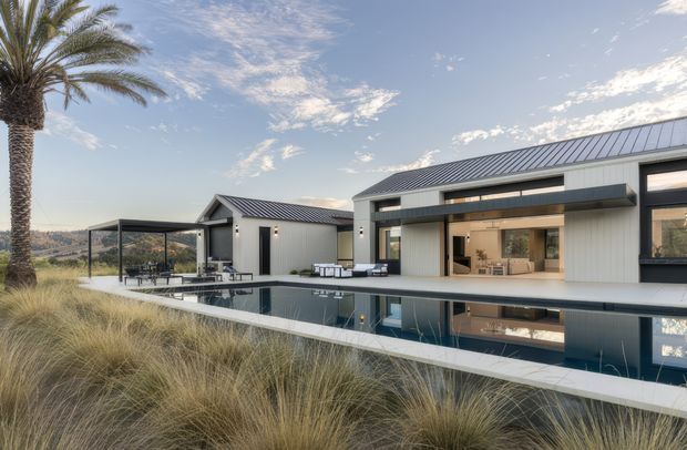 Exterior view of a modern single-storey residence with crisp white and dark grey cladding, low-pitched metal roofs, extensive glazed openings, a covered outdoor dining area, and a long reflecting swimming pool. The house sits within a landscaped, grassy setting with a prominent palm tree and ornamental grasses.
