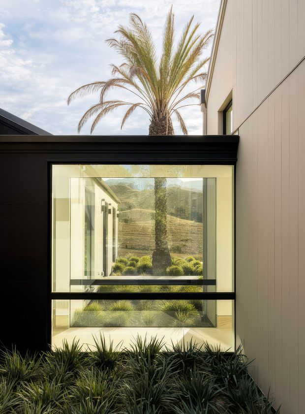 Exterior architectural view of a contemporary house centered on a large black-framed window. The glazing reflects and reveals a landscaped courtyard with grasses, shrubs, a palm tree and a dry, hilly landscape beyond. The building has pale vertical wall cladding, dark roof and window framing, and a carefully framed indoor-outdoor connection.
