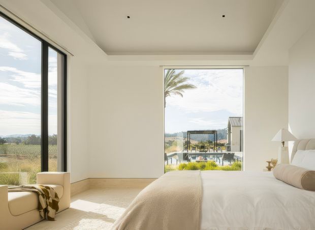 Bright, airy contemporary bedroom with a large upholstered bed, neutral white and cream furnishings, recessed tray ceiling, pale carpet, and expansive black-framed glazing. The windows overlook a landscaped residential setting with a pool and distant hills, while a small armchair and side table occupy the corner.