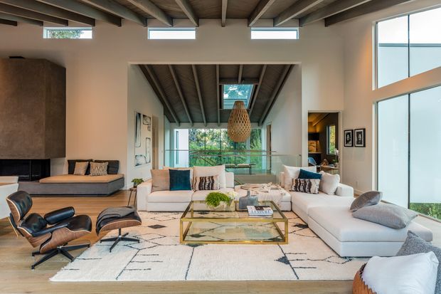 A bright, spacious contemporary living room with exposed timber ceiling beams, extensive floor-to-ceiling glazing, and a glass balustrade overlooking greenery. The room features a large white sectional sofa, neutral textured rug, low glass-and-metal coffee table, sculptural leather lounge chairs, built-in dark cabinetry, and a woven pendant light. A mezzanine or adjoining upper-level space is visible through the central opening.