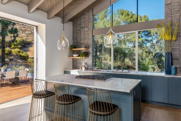 A bright contemporary kitchen with a large stone island and seating for four, dark blue-grey lower cabinetry, integrated cooktop, and pendant lights. Exposed timber ceiling beams and a timber-lined ceiling contrast with textured masonry walls and expansive black-framed glazing. The kitchen opens to a timber outdoor deck and overlooks a leafy, sloping landscape.