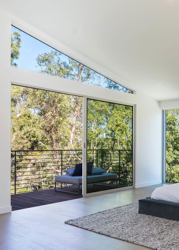 Bright contemporary bedroom with a partially visible low-profile bed, pale wood or tile flooring, and a textured grey area rug. Floor-to-ceiling glazed sliding doors open onto a narrow timber deck with dark horizontal metal balustrading, while high clerestory glazing brings in additional daylight. Dense leafy vegetation is visible outside.