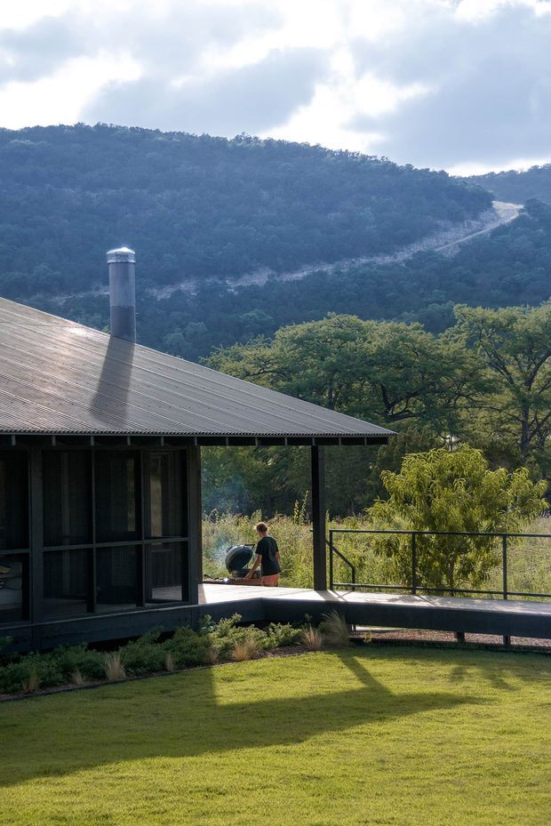 A compact dark-clad cabin or pavilion sits on a landscaped hillside site, with a broad corrugated metal pitched roof, chimney, screened or glazed verandah, and an elevated timber deck with simple black metal railings. The building overlooks dense wooded hills and a dramatic rocky ridgeline, creating a secluded bush setting. A neatly maintained lawn and native-style planting occupy the foreground.