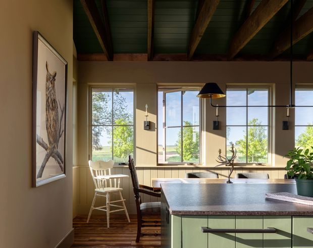 A warm, characterful kitchen and dining area with a large pale green island, dark stone benchtop, timber flooring, and exposed timber beams beneath a dark painted ceiling. Large divided-pane windows bring in abundant natural light and overlook greenery. The space includes a dining table with mismatched timber chairs, black pendant lighting, wall sconces, and a framed bird artwork. The overall feel is rustic and inviting with a strong connection to the outdoors.