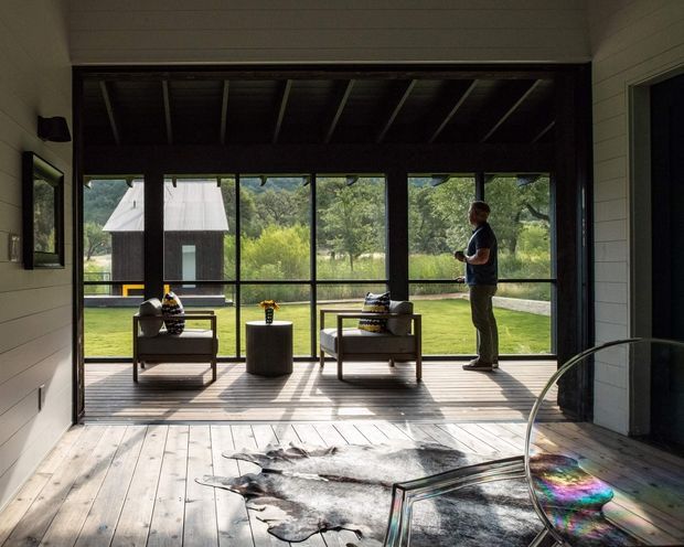 A light-filled enclosed sunroom or indoor-outdoor living space with full-height black-framed glazing overlooking a landscaped lawn and garden. The room has pale horizontal wall cladding, exposed dark ceiling rafters, wide timber floorboards, two upholstered lounge chairs, a small round side table, and a sculptural cowhide rug. A person stands by the glazing, with a dark outbuilding visible beyond.